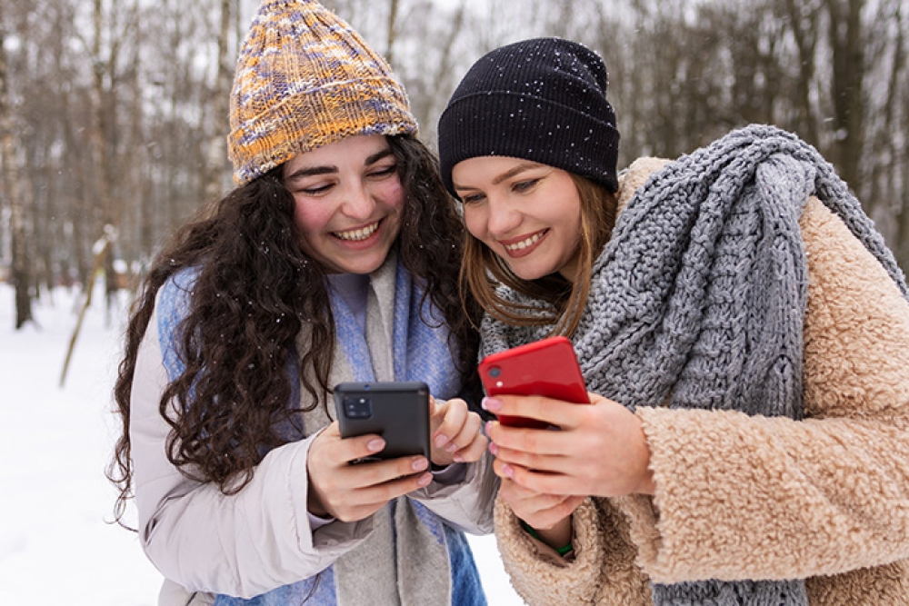 Dwie uśmiechnięte kobiety na śniegu przeglądają aplikację DPD Mobile na swoich smartfonach, planując odbiór przesyłek.